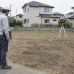 住宅用地の地面を確認する作業着姿の人物と測量用の三脚